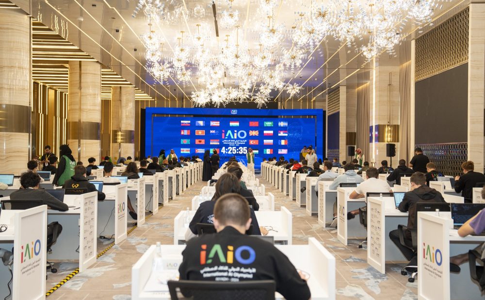 Competitors at individual desks in the AI Olympiad hall, national flags on the stage screen
